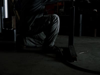 Fabricator kneeling at a steel frame in the open shop bay, the roll-up door light behind and stock racked along the length of the wall.