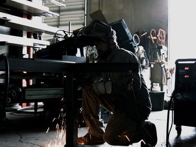 Wide view down the fabrication shop with racked stock to the left, a welder working at a frame, and the roll-up door open to daylight.