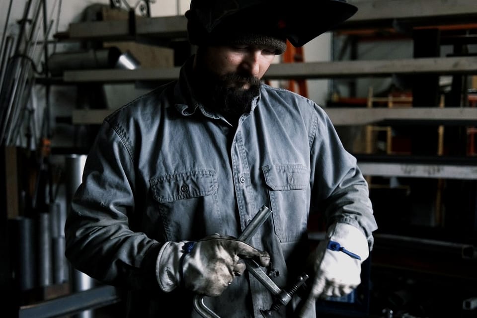 Fabricator in a welding hood and shop coat setting up a cut at the bench, steel stock racked on the wall behind the work area.