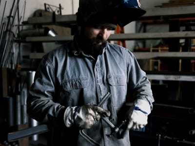 Fabricator in a welding hood and shop coat setting up a cut at the bench, steel stock racked on the wall behind the work area.