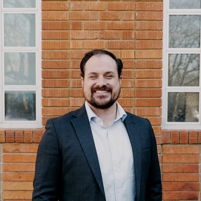 Tyler Miller, founder of Miller 3 Engineering, in a dark blazer and light shirt outside a red brick building with white-framed windows.