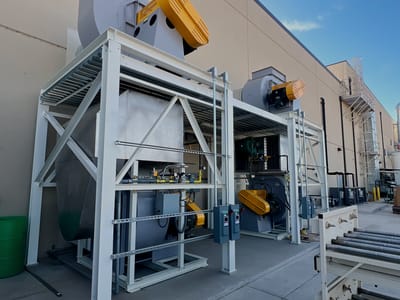 Fabricated collector frame with its blower and drive package hanging from a crane line outside a plant, staged over the receiving apron.