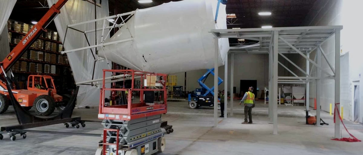 Large white process vessel set on a fabricated steel platform in a warehouse bay, with a scissor lift and a telehandler working beneath it.