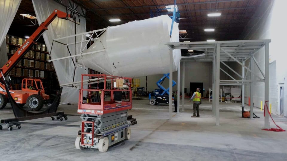 Large white process vessel set on a fabricated steel platform in a warehouse bay, with a scissor lift and a telehandler working beneath it.