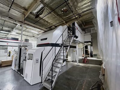 OEM press line set on a plant floor behind temporary containment sheeting, with a fabricated steel access stair and platform on its left flank.