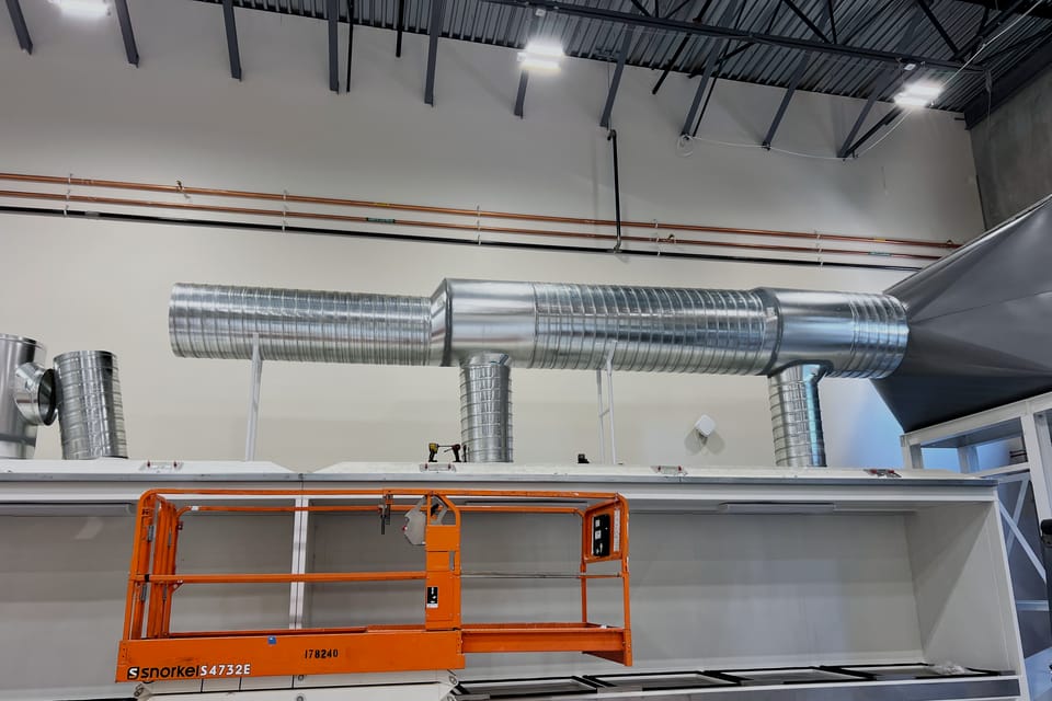 Spiral ductwork branching from a header into the top of a seed dryer body, hung from bar joists above a white powder-coated plenum.
