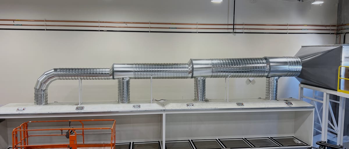 Seed dryer enclosure running the length of a processing bay, with spiral supply ductwork overhead and a scissor lift parked alongside for scale.