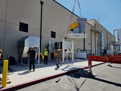 Crew rigging a blower package under a crane line outside a processing plant, with the fabricated frame landed and a bollard in the foreground.