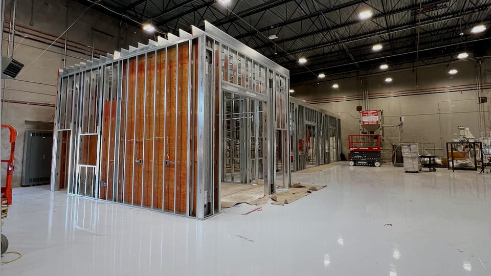Priming room enclosure at framing stage, light-gauge steel studs standing open inside a warehouse bay with a sealed floor and skylights above.
