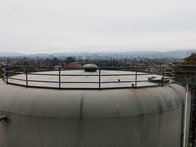 Roof of an in-service industrial water tank with a new perimeter guardrail run around the deck edge, the valley and town below in haze.