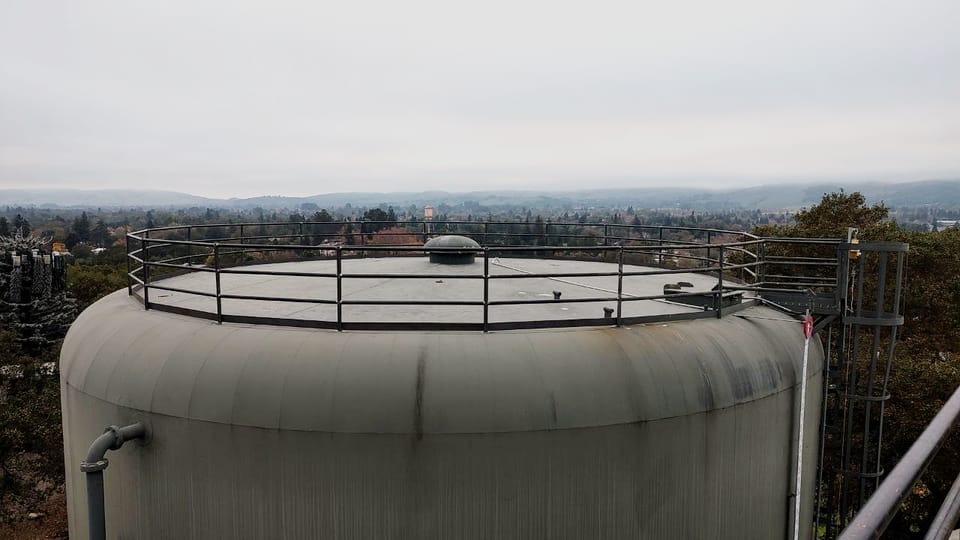 Roof of an in-service industrial water tank with a new perimeter guardrail run around the deck edge, the valley and town below in haze.