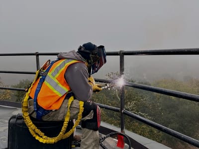 Welder in a harness and high-visibility vest making a field weld at the edge of an elevated tank deck, lanyard tied off to the new rail.