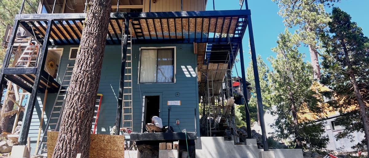 Completed steel deck and balcony wrapping two faces of a hillside house, blackened columns and beams carrying a timber-slatted roof above.