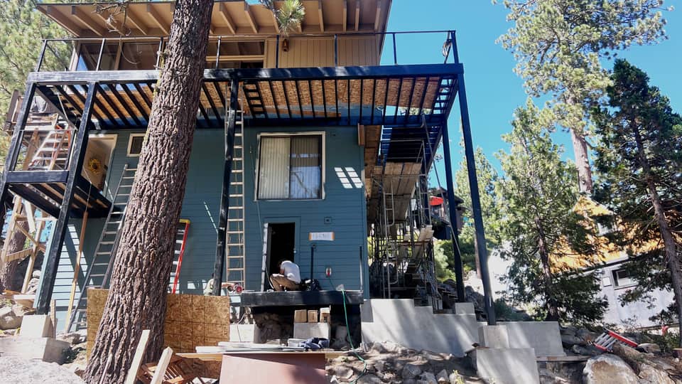 Completed steel deck and balcony wrapping two faces of a hillside house, blackened columns and beams carrying a timber-slatted roof above.