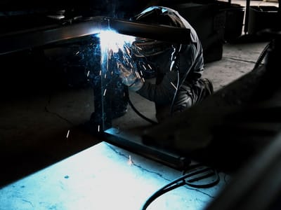 Welder striking an arc on a steel section at a shop bench, the weld pool lighting the table and the hood while the shop behind stays dark.