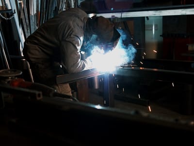 Welder working along a horizontal steel beam laid across the fabrication bay, the arc lighting the flange and web at the joint.
