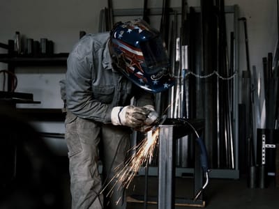 Operator cutting stock at the rack with a hand grinder, sparks streaming down the length of the bar against the racked steel behind.