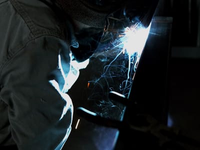 Welder in a hood and leathers running a weld at the shop table, sparks throwing forward across the steel and the arc reflecting off the bench.